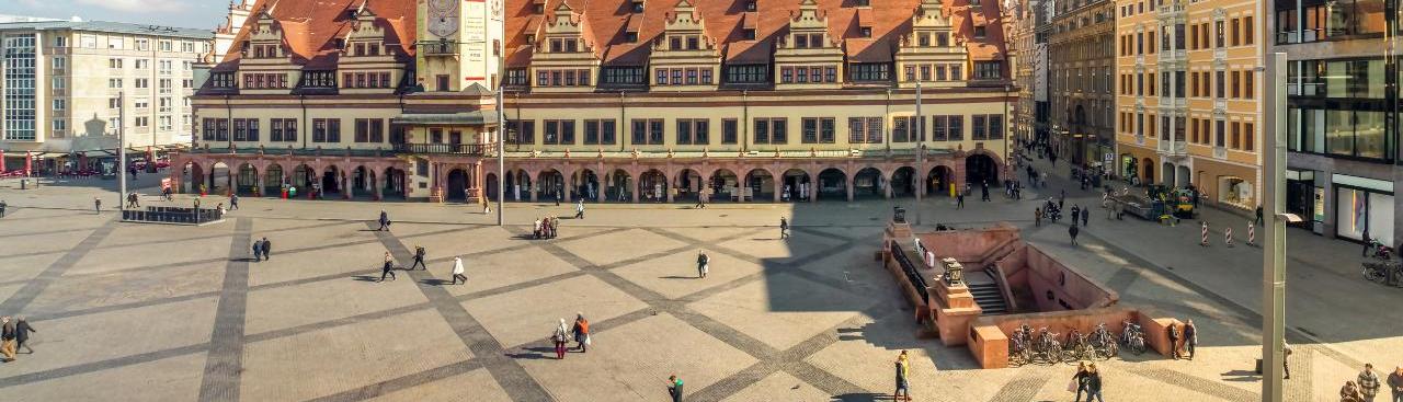 Zu sehen ist der Marktplatz mit dem Alten Rathaus in Leipzig bei Sonnenschein. Verschiedene Personen gehen geschäftig über den sonst leeren Platz.