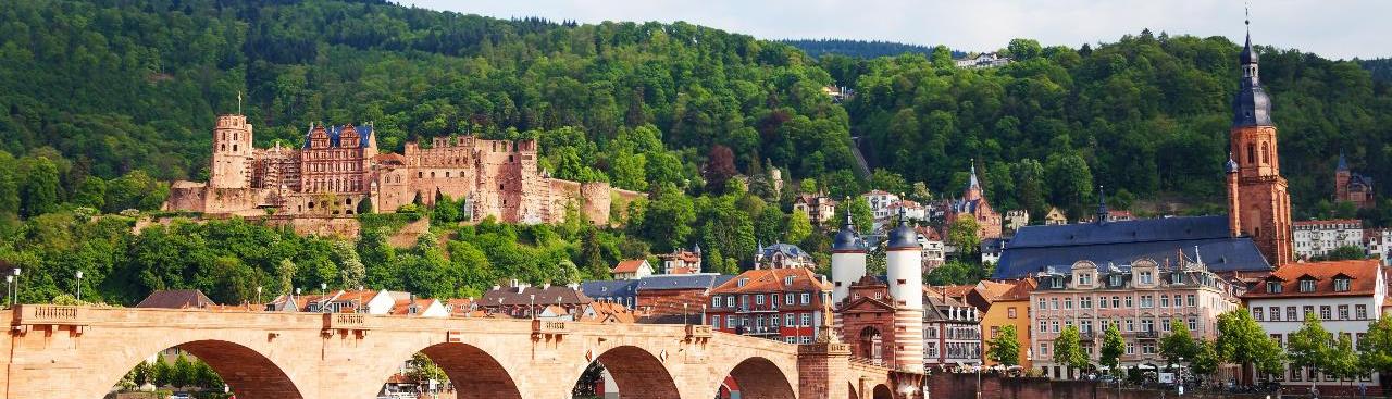 Im Bildvordergrund sieht man den Neckar und die Karl-Theodor-Brücke an einem sonnigen Sommertag. Im Hintergund rechts ist die Altstadt mit einer markanten Kirche und links das Schloss Heidelberg zu sehen.