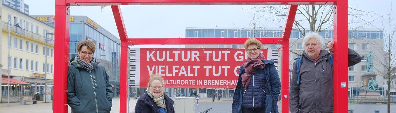 Vier Personen stehen und sitzen unter einer offenen, roten Holzstruktur in einem städtischen Platz, mit dem Banner  Vier Personen stehen und sitzen unter einer offenen, roten Holzstruktur in einem städtischen Platz, mit dem Banner