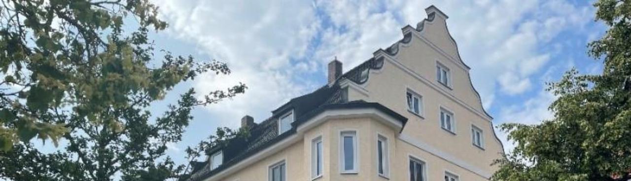 Ein dreistöckiges Eckgebäude mit blassgelber Fassade, weißem Dekor und markantem Spitzdach unter leicht bewölktem Himmel. Ein dreistöckiges Eckgebäude mit blassgelber Fassade, weißem Dekor und markantem Spitzdach unter leicht bewölktem Himmel.