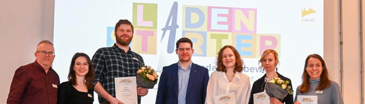 Sieben Personen auf einer Bühne halten Zertifikate und kleine Blumensträuße. Im Hintergrund eine Präsentation auf einer Leinwand mit einem Banner  Sieben Personen auf einer Bühne halten Zertifikate und kleine Blumensträuße. Im Hintergrund eine Präsentation auf einer Leinwand mit einem Banner