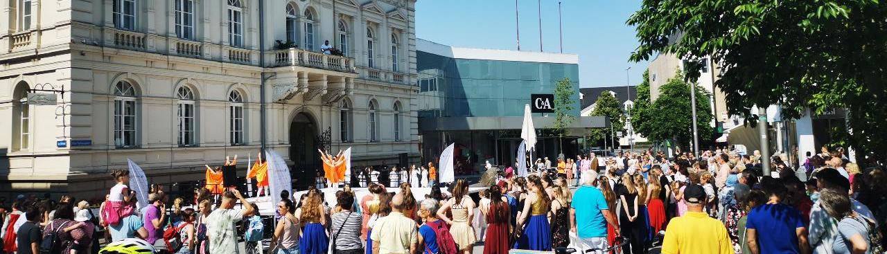 Ein sonniger Tag mit einer großen Menschenmenge im Freien vor einem klassischen Gebäude mit Uhrturm und modernen Strukturen im Hintergrund. Auf einer Bühne tanzen Menschen in orangefarbenen Gewändern. Ein sonniger Tag mit einer großen Menschenmenge im Freien vor einem klassischen Gebäude mit Uhrturm und modernen Strukturen im Hintergrund. Auf einer Bühne tanzen Menschen in orangefarbenen Gewändern.