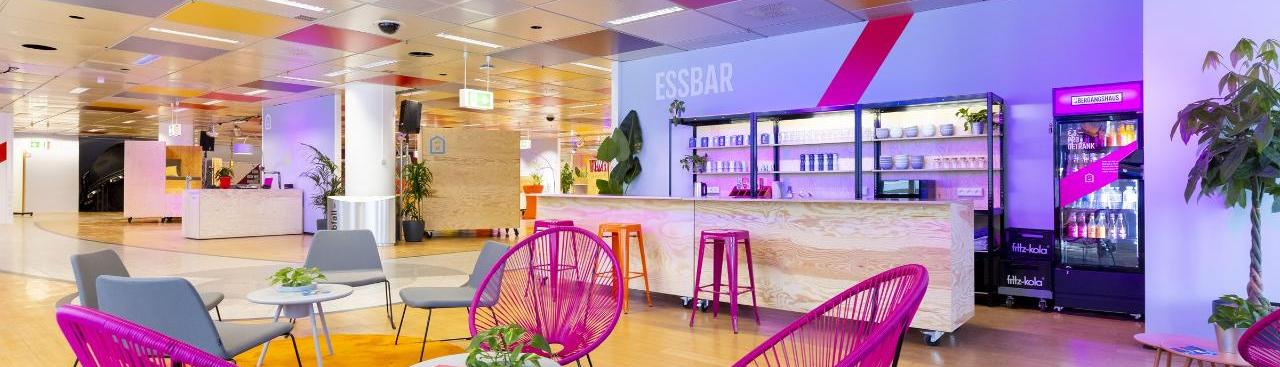 Farbenfroher Innenraum mit pink-, grau- und orangefarbenen Stühlen und Teppichen und einer beleuchteten Holzbar mit der weißen Aufschrift  Farbenfroher Innenraum mit pink-, grau- und orangefarbenen Stühlen und Teppichen und einer beleuchteten Holzbar mit der weißen Aufschrift