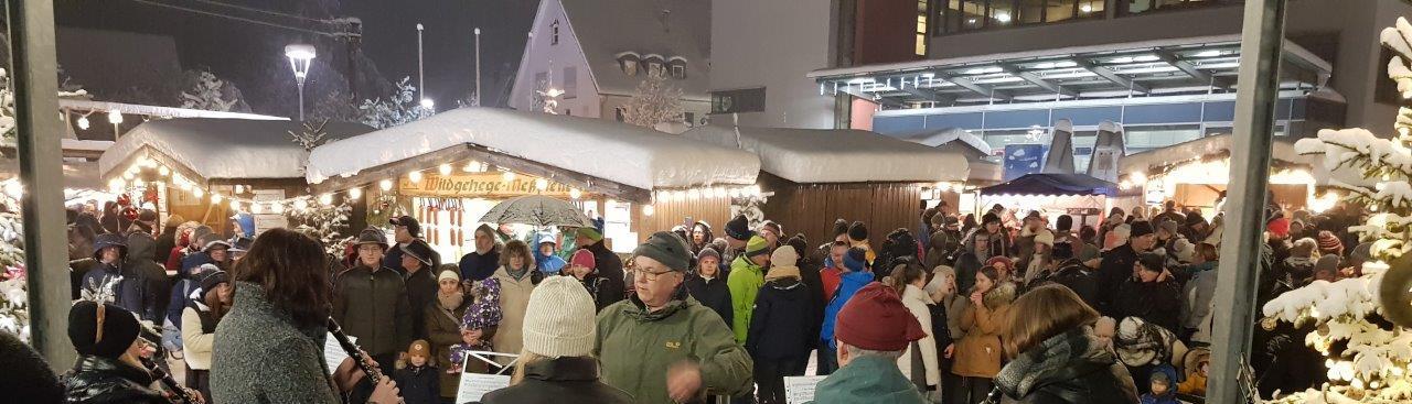 Ein belebter Weihnachtsmarkt im Freien mit zahlreichen Besuchern und Besucherinnen, die einer Musikgruppe in der Mitte zuhören. Die Szene ist mit Dekoration und Lichtern geschmückt. Im Hintergrund sind Marktstände. Ein belebter Weihnachtsmarkt im Freien mit zahlreichen Besuchern und Besucherinnen, die einer Musikgruppe in der Mitte zuhören. Die Szene ist mit Dekoration und Lichtern geschmückt. Im Hintergrund sind Marktstände.
