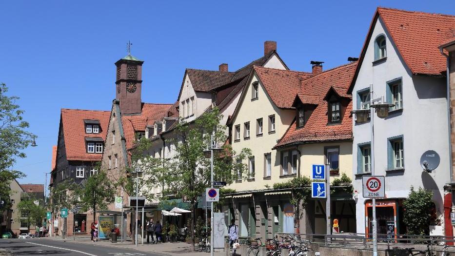 Auf dem Fußweg einer Einkauffsstraße in Fürth mit historischer Fassade laufen einzelen Passanten. Der Gehweg ist von grünen Bäumen gesäumt und mittig befindet sich eine Bushaltestelle. Vorne rechts sind der Abgang zu einer Unterführung und Fahrräder.