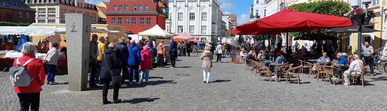 In Greifswald findet ein Markt unter blauem Himmel statt. Rechts im Bild sitzen Café Gäste unter roten Sonnenschirmen