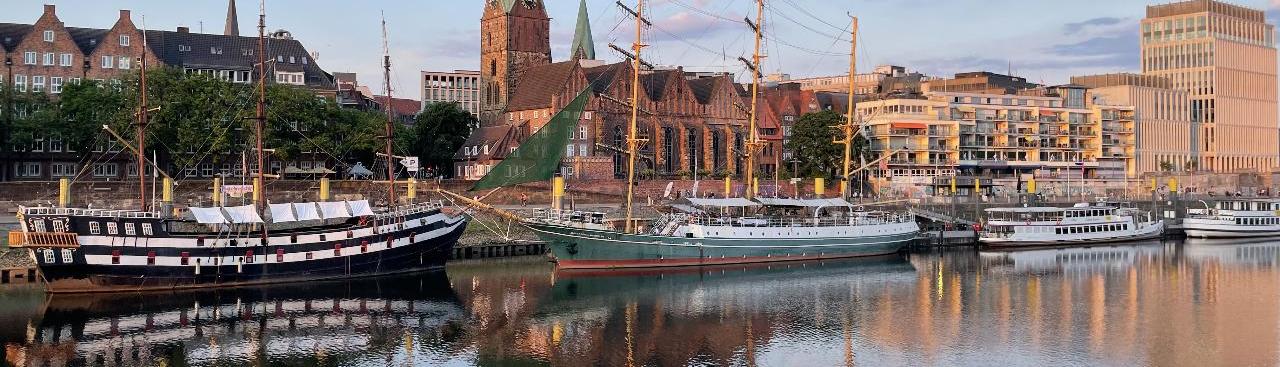 In einem Hafen liegen zwei historische Segelschiffe und zwei Boote am Kai. Dahinter stehen links Häuser hinter Bäumen, mittig eine Kirche aus rotem Backstein und rechts modernere helle Gebäude. Die Boote spiegeln sich im Wasser, die Sonne geht unter. In einem Hafen liegen zwei historische Segelschiffe und zwei Boote am Kai. Dahinter stehen links Häuser hinter Bäumen, mittig eine Kirche aus rotem Backstein und rechts modernere helle Gebäude. Die Boote spiegeln sich im Wasser, die Sonne geht unter.