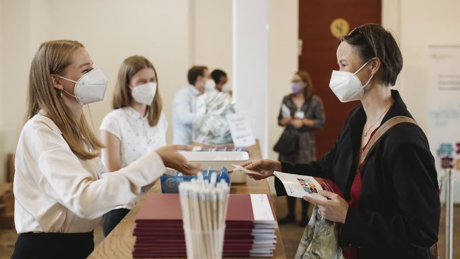 Über einen Tresen hinweg reicht eine Frau einer anderen Frau ein Namensschild. Im Hintergrund stehen weitere Personen. Auf dem Tresen liegen weinrote Tagungsmappen und steht ein Glas mit Bleistiften. Alle Anwesenden tragen weiße FFP2-Masken. Über einen Tresen hinweg reicht eine Frau einer anderen Frau ein Namensschild. Im Hintergrund stehen weitere Personen. Auf dem Tresen liegen weinrote Tagungsmappen und steht ein Glas mit Bleistiften. Alle Anwesenden tragen weiße FFP2-Masken.