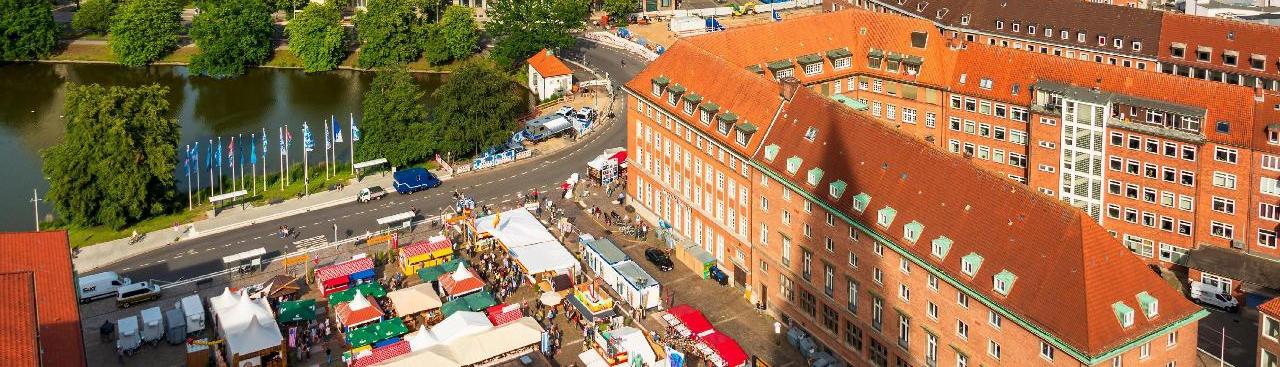 Ein Luftbild der Innenstadt Kiels bei Sonnenschein im Sommer. Im Vordergrund links sieht man einen Markt mit Ständen und Menschen, dahinter einen See mit Bäumen. Hinter den Gebäuden der Innenstadt fällt der Blick auf den Hafen mit einem Kran und Schiffen