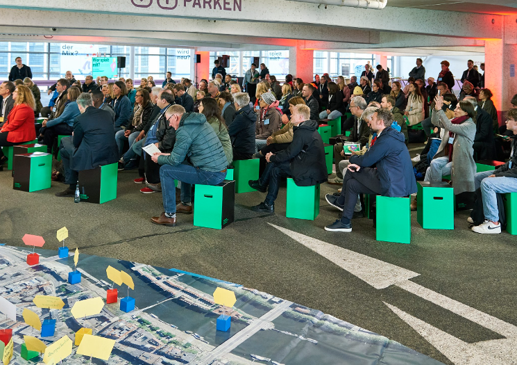 Menschen sitzen auf grünen Würfeln in einem Parkhaus, daneben ein Stadtmodell auf dem Boden. Menschen sitzen auf grünen Würfeln in einem Parkhaus, daneben ein Stadtmodell auf dem Boden.