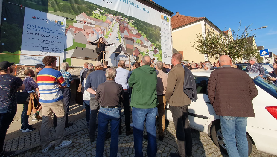 Auf einer Bühne steht eine dunkel gekleidete Frau mit geöffneten Armen. Die Sonne scheint. Davor stehen stehen viele Menschen und schauen in Richtung Bühne. Die Menschen stehen um zwei Autos herum. Neben der Bühne steht ein Baum. Auf einer Bühne steht eine dunkel gekleidete Frau mit geöffneten Armen. Die Sonne scheint. Davor stehen stehen viele Menschen und schauen in Richtung Bühne. Die Menschen stehen um zwei Autos herum. Neben der Bühne steht ein Baum.