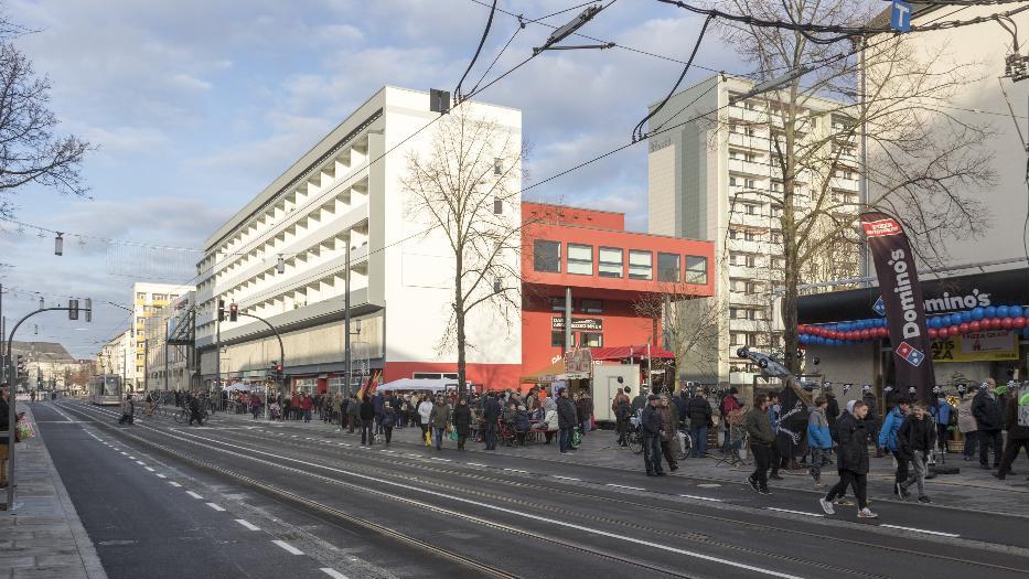Auf dem Gehweg neben einer Hauptstraße mit Straßenbahnschienen gehen viele Menschen. Über der Straße hängt eine Oberleitung. Dahinter stehen mehrgeschossige Plattenbauten in weiß, ein Gebäude ist orange gestrichen. Davor wachsen große Bäume, ohne Blätter. Auf dem Gehweg neben einer Hauptstraße mit Straßenbahnschienen gehen viele Menschen. Über der Straße hängt eine Oberleitung. Dahinter stehen mehrgeschossige Plattenbauten in weiß, ein Gebäude ist orange gestrichen. Davor wachsen große Bäume, ohne Blätter.