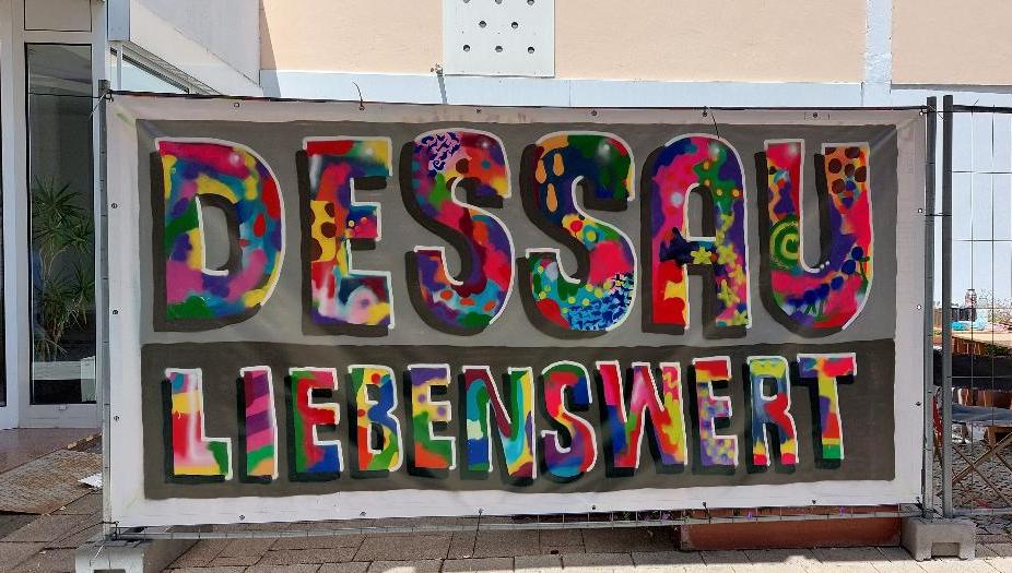 Auf einer Plane an einem Bauzaun ist der Slogan "Dessau liebenswert" gut leserlich in sehr bunten Buchstaben als Graffiti gesprayt. Auf einer Plane an einem Bauzaun ist der Slogan "Dessau liebenswert" gut leserlich in sehr bunten Buchstaben als Graffiti gesprayt.