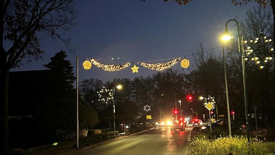Über einer Straße und an Laternen ist an mehreren Stellen Winterbeleuchtung in Form von Sternen und Leuchtgirlanden gespannt. Diese und die Laternen beleuchten einen dunklen Abend im Dezemeber. Neben der Straße stehen Bäume, weiter hinten Autos. Über einer Straße und an Laternen ist an mehreren Stellen Winterbeleuchtung in Form von Sternen und Leuchtgirlanden gespannt. Diese und die Laternen beleuchten einen dunklen Abend im Dezemeber. Neben der Straße stehen Bäume, weiter hinten Autos.