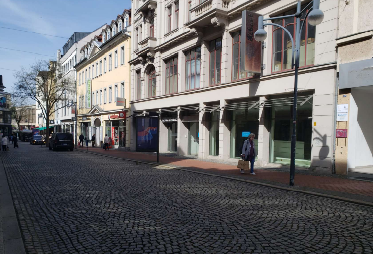 An einer Kopfsteinpflasterstraße stehen mehre gründerzeitliche Häuser mit Geschäften im Erdgeschoss. Davor geht eine Frau, im Hintergrund steht ein einzelner Baum ohne Blätter und mehrere Autos. Die Sonne scheint. An einer Kopfsteinpflasterstraße stehen mehre gründerzeitliche Häuser mit Geschäften im Erdgeschoss. Davor geht eine Frau, im Hintergrund steht ein einzelner Baum ohne Blätter und mehrere Autos. Die Sonne scheint.