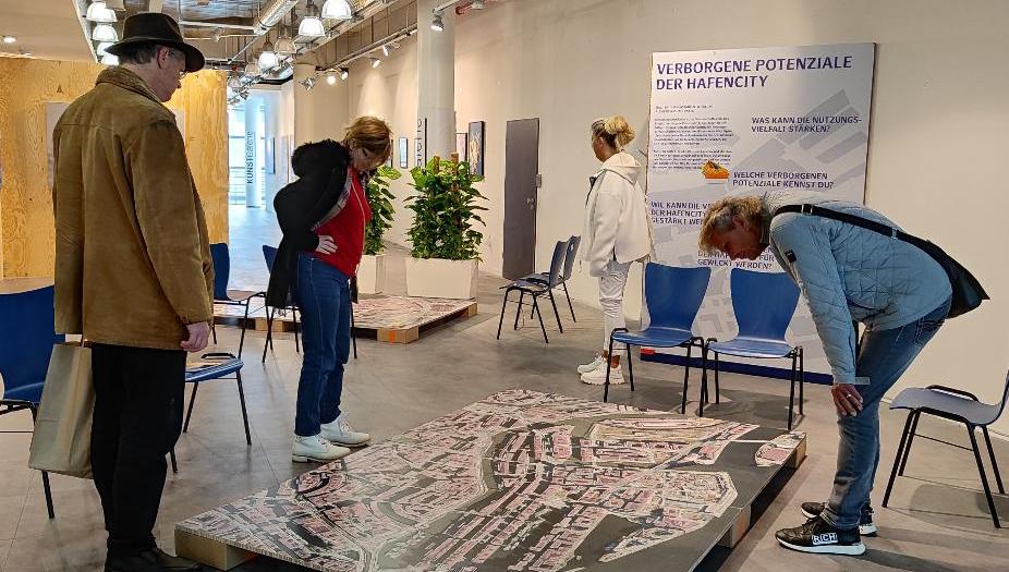 Zwei Frauen und ein Mann mit Hut betrachten ein gedrucktes großes Luftbild, das auf einer Palette in einem Raum liegt. Daneben stehen zwei blaue Stühle, dahinter ein Roll-Up mit der Überschrift: "Verborgene Potential Hafen-City Hamburg". 