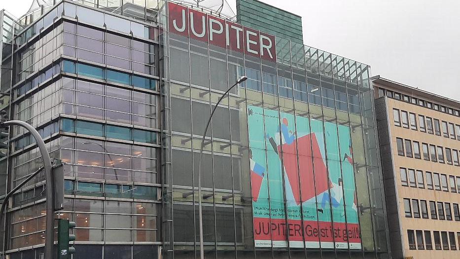 In einem viergeschössigen Gebäude mit Glasfassade zur Straße hängt ein großes Plakat mit  derabstrakten Darstellung zweier Personen, die ein rotes Qaudrat tragen. Darunter steht "Jupiter. Geist ist Geil." Im obersten Stock steht erneut "Jupiter".