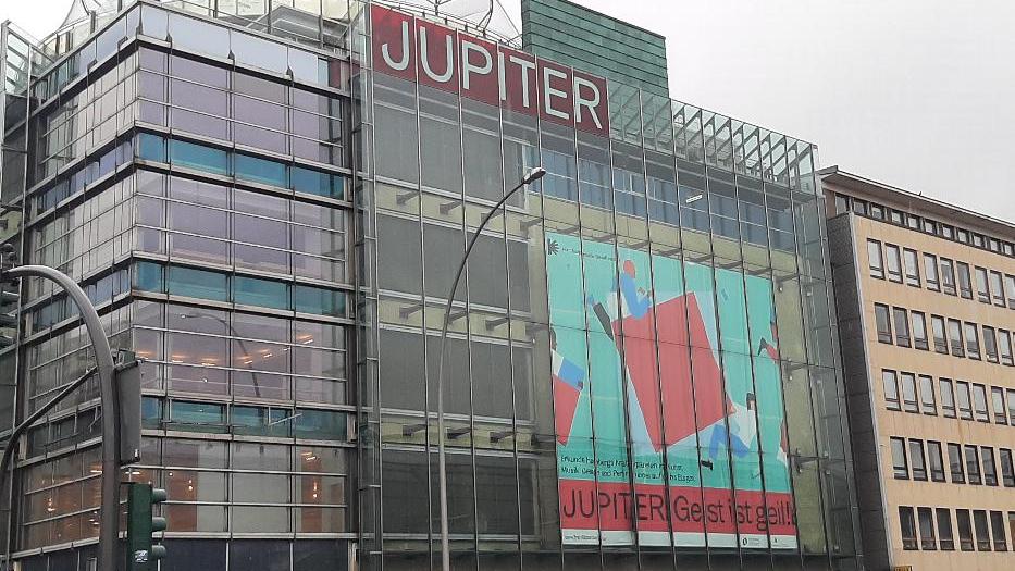 In einem viergeschössigen Gebäude mit Glasfassade zur Straße hängt ein großes Plakat mit derabstrakten Darstellung zweier Personen, die ein rotes Qaudrat tragen. Darunter steht "Jupiter. Geist ist Geil." Im obersten Stock steht erneut "Jupiter". In einem viergeschössigen Gebäude mit Glasfassade zur Straße hängt ein großes Plakat mit derabstrakten Darstellung zweier Personen, die ein rotes Qaudrat tragen. Darunter steht "Jupiter. Geist ist Geil." Im obersten Stock steht erneut "Jupiter".