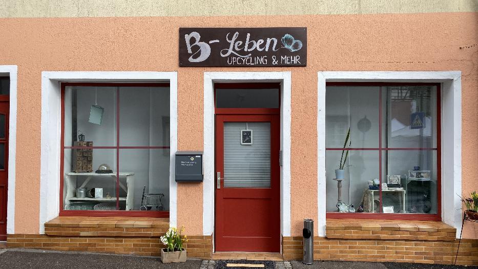 Über der roten Tür eines Ladenlokals steht "B-Leben, Upcycling & mehr" in Handschrift. Die Wand des Gebäudes ist blass oragne, die Fensterbänke braun geklinkert. In den rot gerahmten Schaufenstern stehen viele kleine Gegenstände und ein kleiner Kaktus. Über der roten Tür eines Ladenlokals steht "B-Leben, Upcycling & mehr" in Handschrift. Die Wand des Gebäudes ist blass oragne, die Fensterbänke braun geklinkert. In den rot gerahmten Schaufenstern stehen viele kleine Gegenstände und ein kleiner Kaktus.