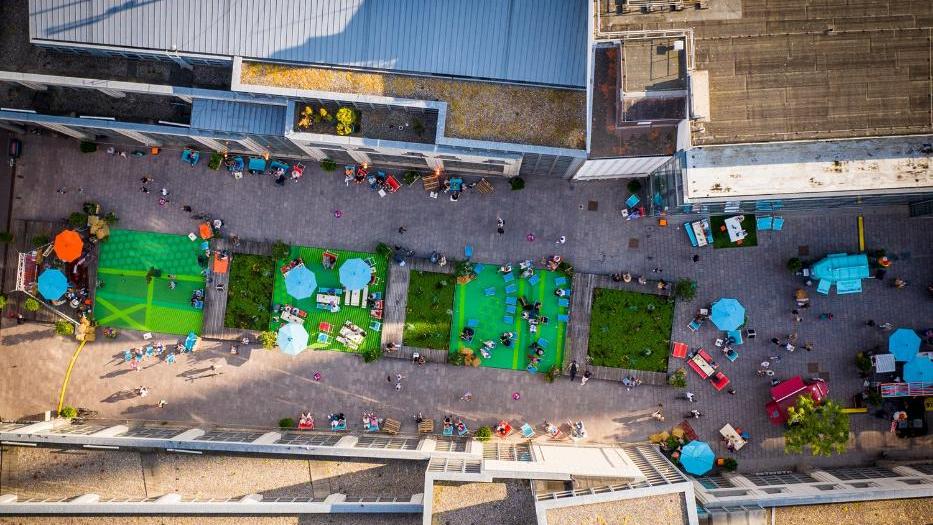 Ein lebendiger Stadtpark aus der Vogelperspektive, umgeben von Gebäuden. Grüne, quadratische Rasenflächen und bunte Spielgeräte mit Menschen, die den Park genießen. Ein lebendiger Stadtpark aus der Vogelperspektive, umgeben von Gebäuden. Grüne, quadratische Rasenflächen und bunte Spielgeräte mit Menschen, die den Park genießen.