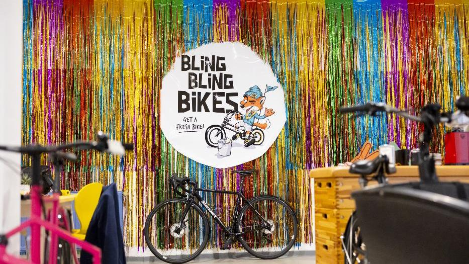Ein farbenfroher Raum gefüllt mit Fahrrädern und einer bunten Lametta-Kulisse im Hintergrund. Im Zentrum ein Schild "BLING BLING BIKES" und ein Fuchs auf einem Fahrrad. Die Atmosphäre wirkt einladend. Ein farbenfroher Raum gefüllt mit Fahrrädern und einer bunten Lametta-Kulisse im Hintergrund. Im Zentrum ein Schild "BLING BLING BIKES" und ein Fuchs auf einem Fahrrad. Die Atmosphäre wirkt einladend.