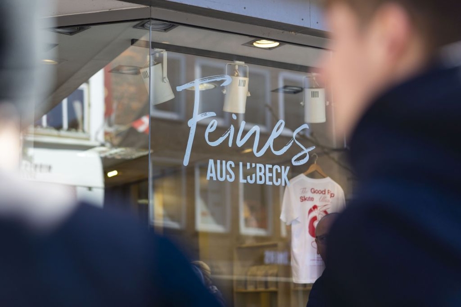 Die Fensterscheibe eines Ladens steht "Feines aus Lübeck". Im Laden selbst ist Licht an und vor der Kamera laufen Menschen verschwommen durch das Bild. Die Fensterscheibe eines Ladens steht "Feines aus Lübeck". Im Laden selbst ist Licht an und vor der Kamera laufen Menschen verschwommen durch das Bild.