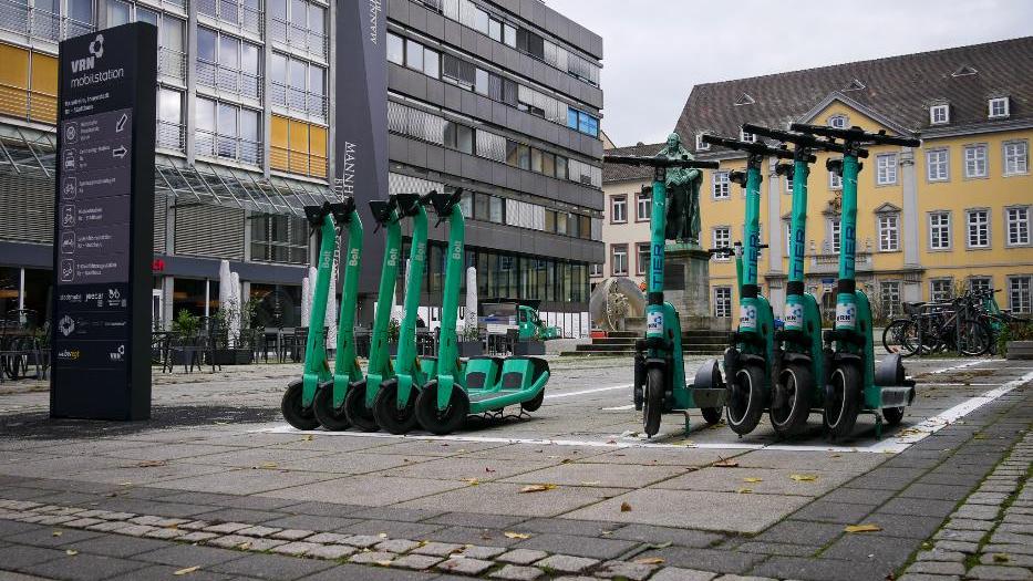 Auf einem Platz in einer Stadt stehen mehrere grüne E-Tretroller in einem weißen aufgemalten Kasten, dahinter viele Fahrräder. Links daneben steht eine Informationstafel in grau mit der Überschrift "VRN-Mobilstation". Im Hintergrund steht eine Skulptur.