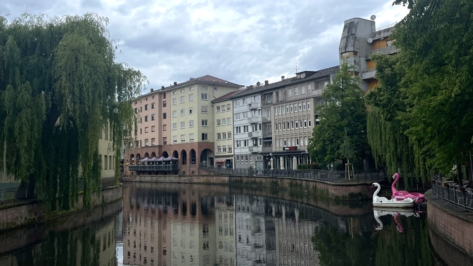 Ein Kanal mit Gebäuden auf einer Seite und Bäumen auf der anderen. Zwei schwanenförmige Tretboote, eines weiß und eines rosa, sind am Kanal angedockt. Die gebäude spiegeln sich im Wasser. Der Tag scheint bewölkt und regnerisch. Ein Kanal mit Gebäuden auf einer Seite und Bäumen auf der anderen. Zwei schwanenförmige Tretboote, eines weiß und eines rosa, sind am Kanal angedockt. Die gebäude spiegeln sich im Wasser. Der Tag scheint bewölkt und regnerisch.