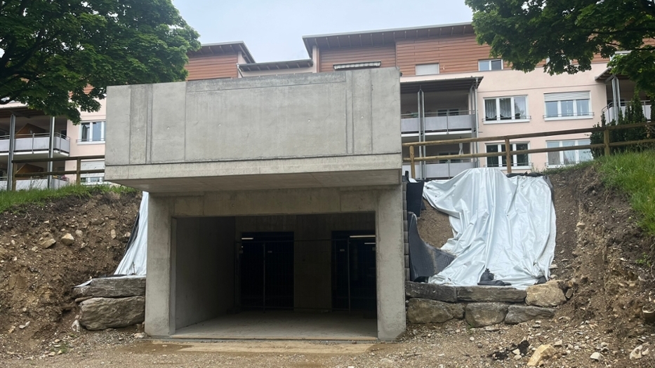 Ein neu gebauter massiver Betonunterstand, der unter einer Straße verläuft und als Zugang zum Parkhaus fungiert. Auf beiden Seiten ist der Eingang von großen Steinen und Bäumen umgeben. Ein neu gebauter massiver Betonunterstand, der unter einer Straße verläuft und als Zugang zum Parkhaus fungiert. Auf beiden Seiten ist der Eingang von großen Steinen und Bäumen umgeben.