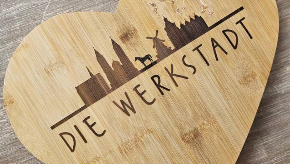 Auf einem Holzbrett ist die zweidimensionale Silouette mehrer Gebäude, darunter eine Kirche und eine Windmühle, sowie eines Pferds eingebrannt. Darunter steht in verspielten Druckbuchstaben "DIE WERKSTATT". Auf einem Holzbrett ist die zweidimensionale Silouette mehrer Gebäude, darunter eine Kirche und eine Windmühle, sowie eines Pferds eingebrannt. Darunter steht in verspielten Druckbuchstaben "DIE WERKSTATT".