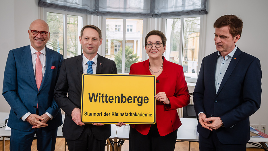 Vier Personen in Anzügen und Kostüm präsentieren ein gelbes Stadtschild mit der Aufschrift "Wittenberge - Standort der Kleinstadtakademie" in einem hellen Raum.