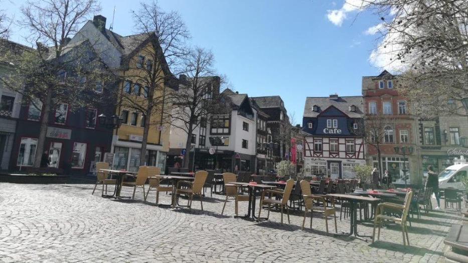 Auf einem Kopfsteinpflasterplatz stehen viele Tische und Stühle eines Cafés, an denen niemand sitzt. Umrahmt ist der Platz mit Bäumen, an denen die ersten Blätter sprießen, dahinter sieht man die historischen Häuser einer Altstadt. Die Sonne scheint.