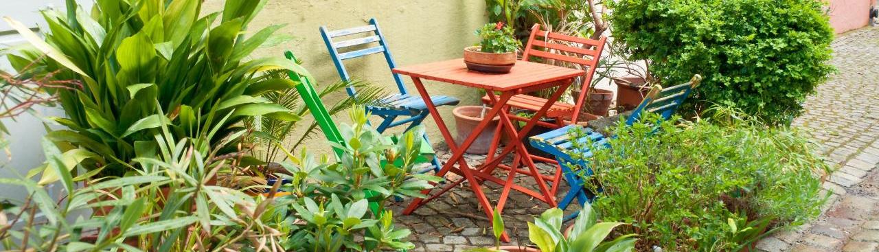 In der Lindauer Altstadt steht ein kleiner Tisch mit Stühlen auf Kopfsteinpflaster. Die Sitzecke ist von vielen Pflanztöpfen mit grünen Büschen und Sträuchern gerahmt. Hinter der Sitzecke sieht man die Häuser der Altstadt mit Fensterläden